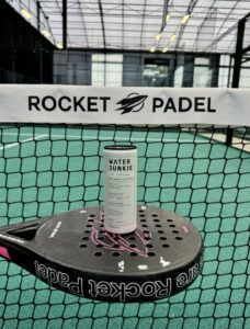 Water Junkie Apple and Elderflower can on Padel racket in front of Rocket Padel net
