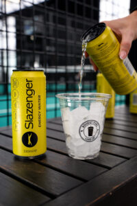 Slazenger Energy Water can standing on table, facing the camera next to to a cup filled with ice. A hand is pouring a can of Slazenger Energy Water into the cup over ice.