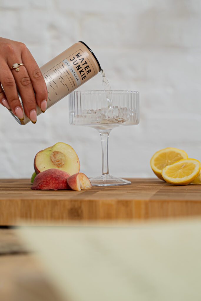 A hand pouring Water Junkie White Peach and Ginger flavour into a glass on a wooden surface with peaches either side