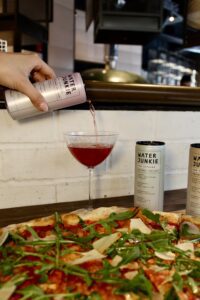 A hand pouring a can of Water Junkie Wild Berry flavour into a glass next to a Homeslice pizza and other Water Junkie flavours