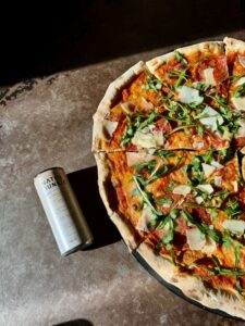 Water Junkie White Peach and Ginger can laying on a table next to a Homeslice pizza with the label facing upwards